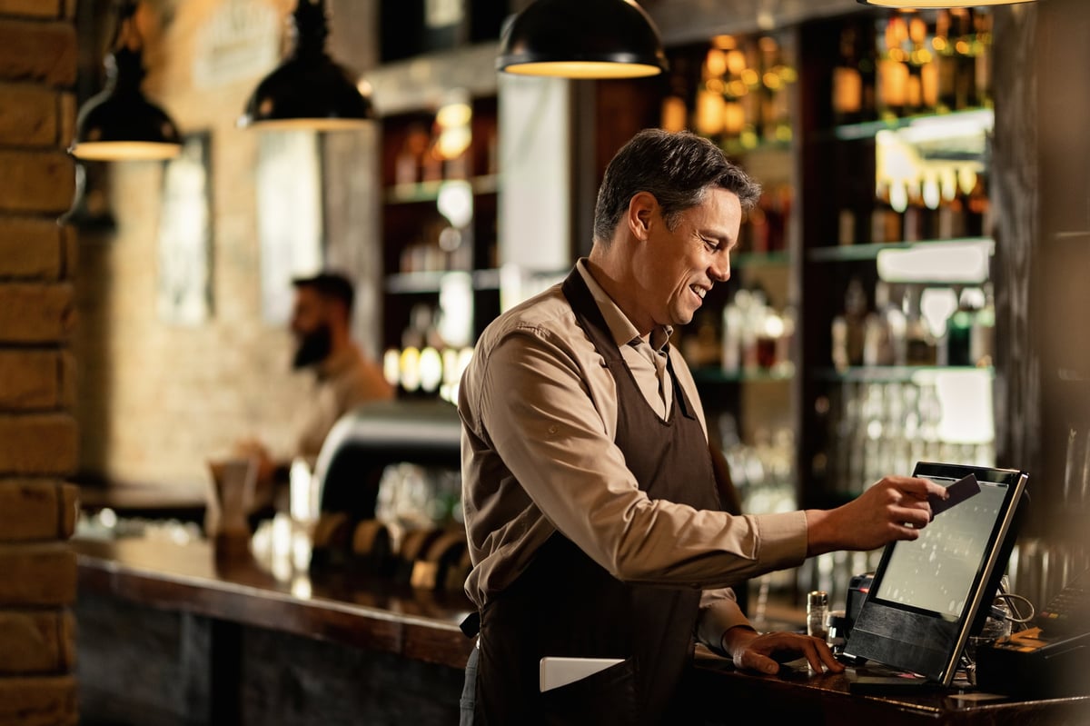 Male bartender in brown apron operating a payment terminal at a bar counter with warm pendant lighting